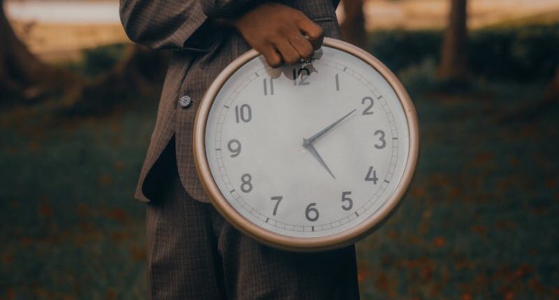 Man in a brown suit outdoors holding a large clock, deep in thought.