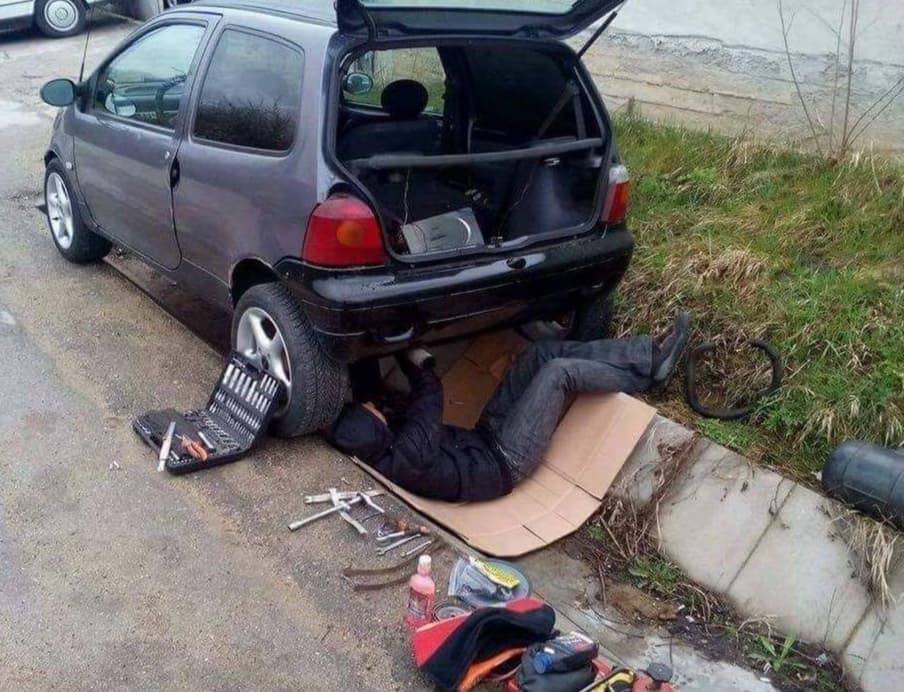 Človek leží na kartóne pri zadnej časti auta vedľa obrubníka a rozloženého náradia.