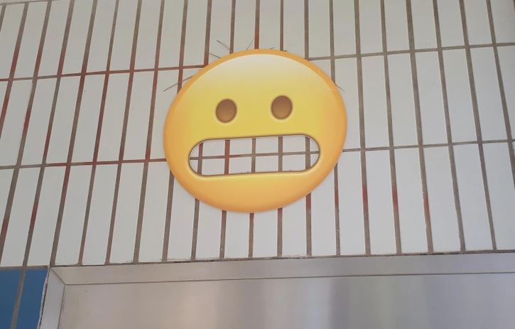 Emoji s nervóznym výrazom na mriežkovanej stene.