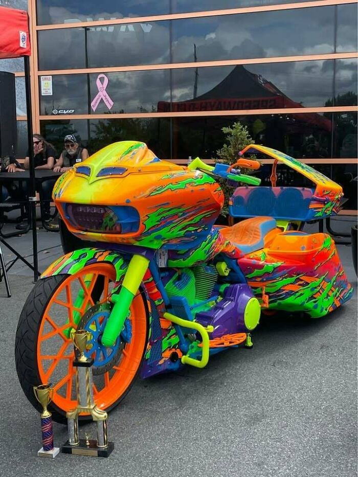 Pestrofarebná motorka s trofejami, parkovaná pred modernou budovou s odrazom čiernej strechy na presklenej fasáde.