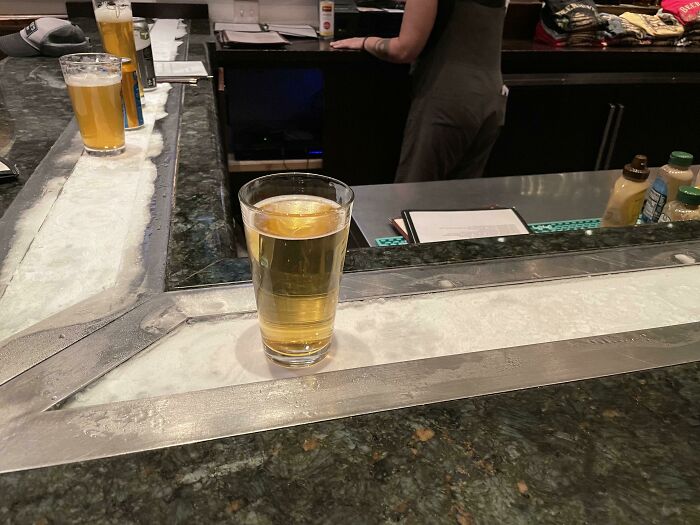 Pohár piva na barovom pulte s ľadovou vrstvou, za ním barmanka.