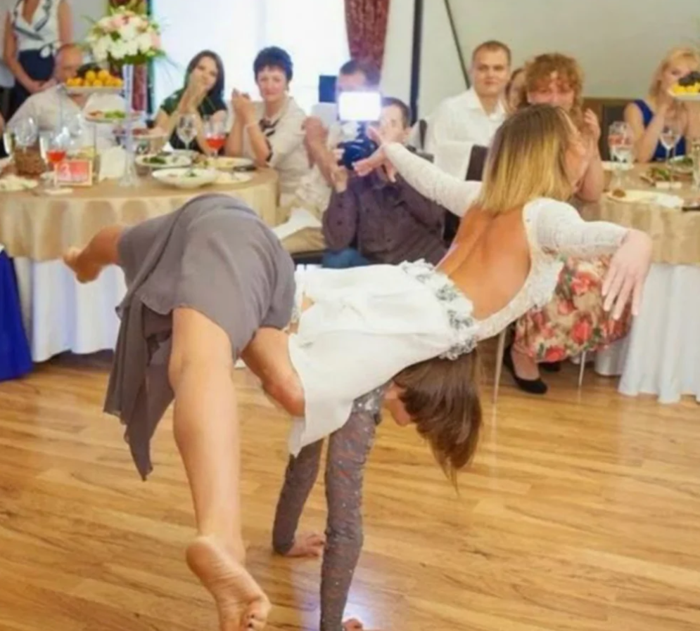 Žena v bielych šatách vykonáva akrobatický tanec na svadbe, pritom ju fotografuje hosť. Publikum aplauzuje.