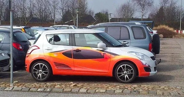 Farebný hatchback s výraznou grafickou úpravou zaparkovaný na parkovisku, okolo iné vozidlá a stromy v pozadí.