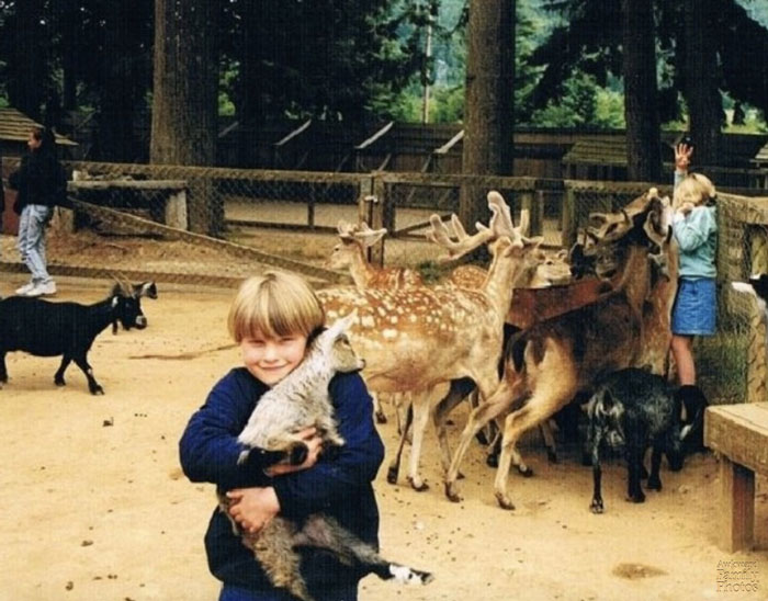 Dieťa objíma kozu v parku so zvieratami, zatiaľ čo ďalšie deti interagujú s jeleňmi a kozami v pozadí.