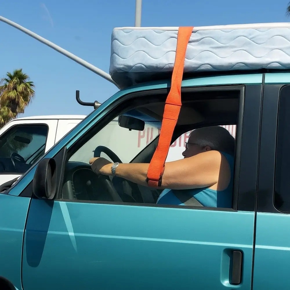 Jazdí auto s matracom pripevneným na streche oranžovým popruhom, modrá dodávka, palma v pozadí.