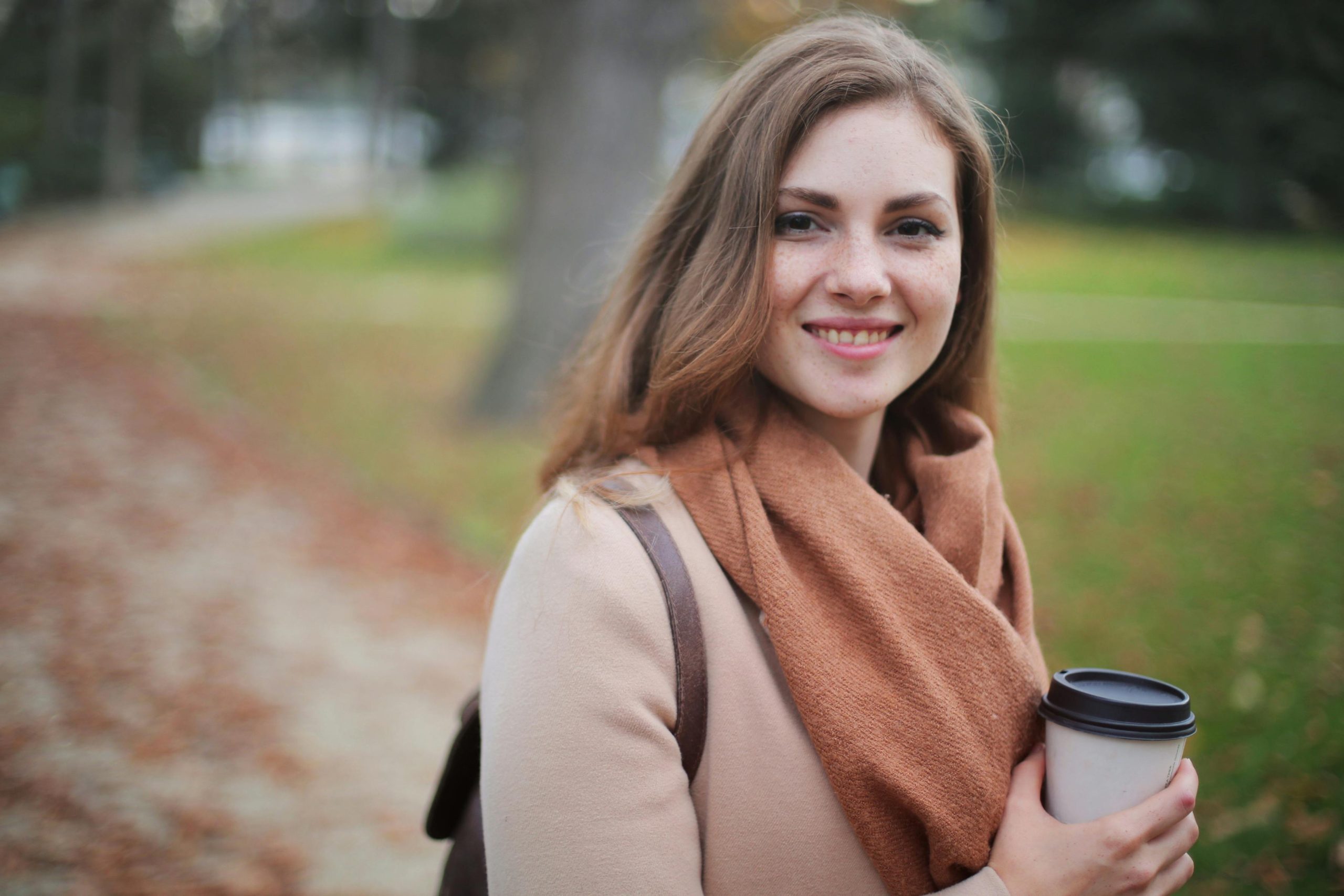 Žena v parku s kávou v ruke, usmievajúc sa, oblečená v jesennom šále a kabáte, rozmazané pozadie.
