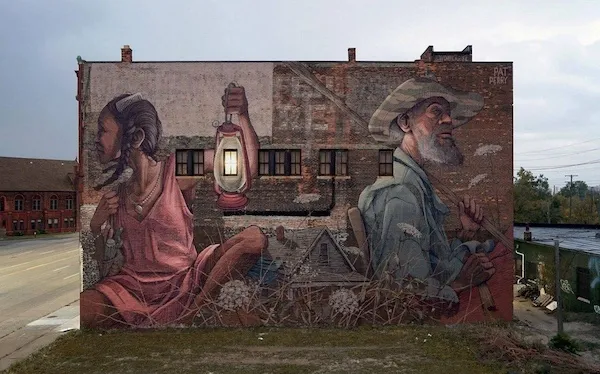 Mural na tehlovej stene s mužom, ženou a petrolejovou lampou, vytvárajúci historickú atmosféru v mestskom prostredí.