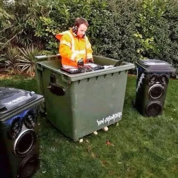 DJ v oranžovej veste hrajúci na DJ pult v kontajneri medzi reproduktormi na tráve. Kreatívna hudobná scéna vonku.