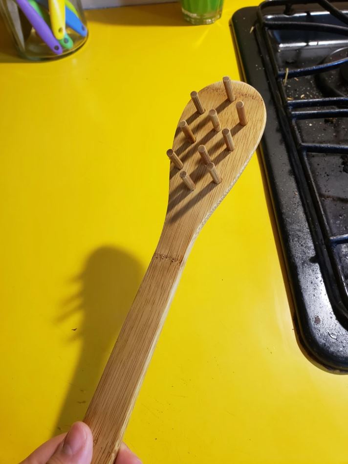 Drevená kuchynská lyžica s kolíkmi na žltom stole vedľa sporáka.