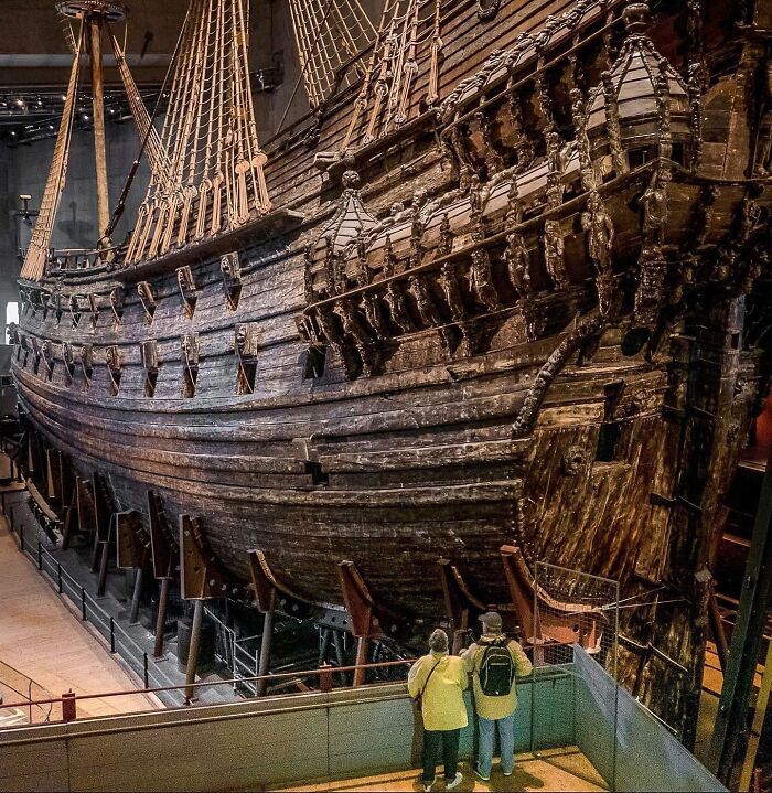 Historická drevená loď v múzeu, návštevníci si prezerajú detaily konštrukcie a výzdoby trupu.