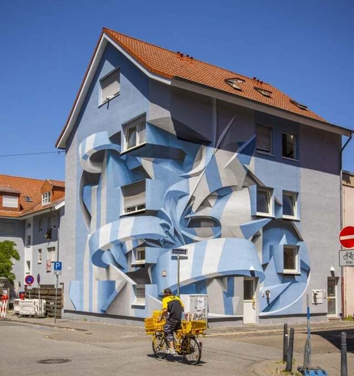 Modrá budova so živým 3D graffiti umením v ulici, kút s cyklistom v žltej veste.