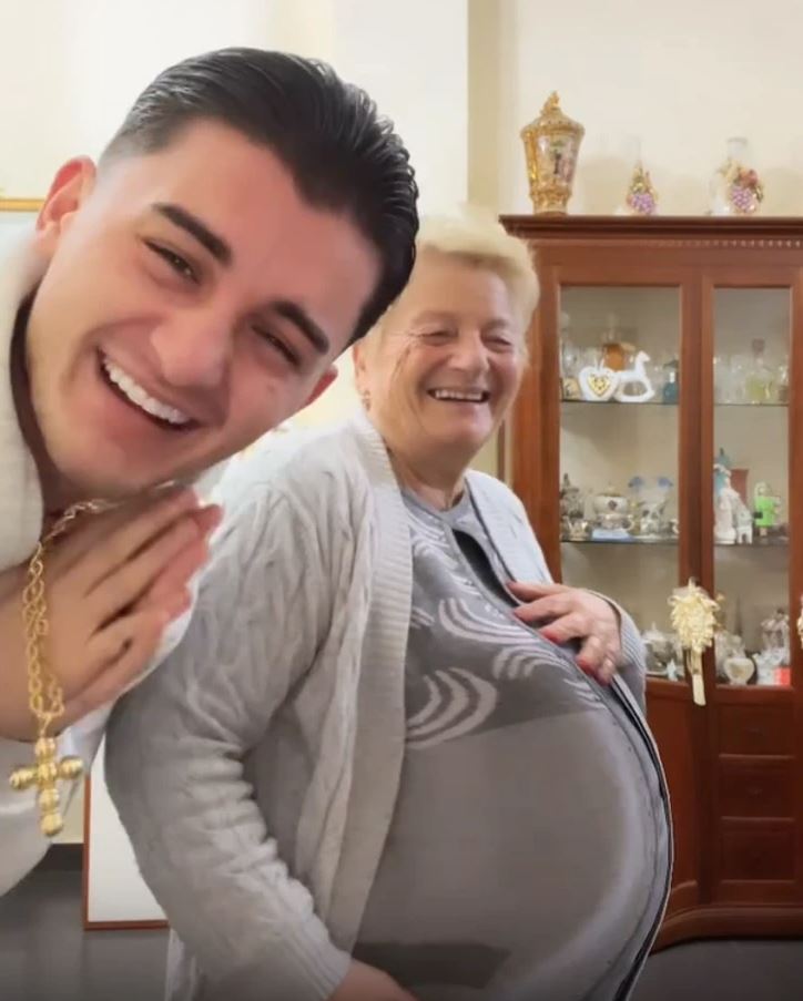Osoby sa smejú v obývačke, žena drží veľký balón pod svetrom, zatiaľ čo muž sa veselo pozerá na fotoaparát.