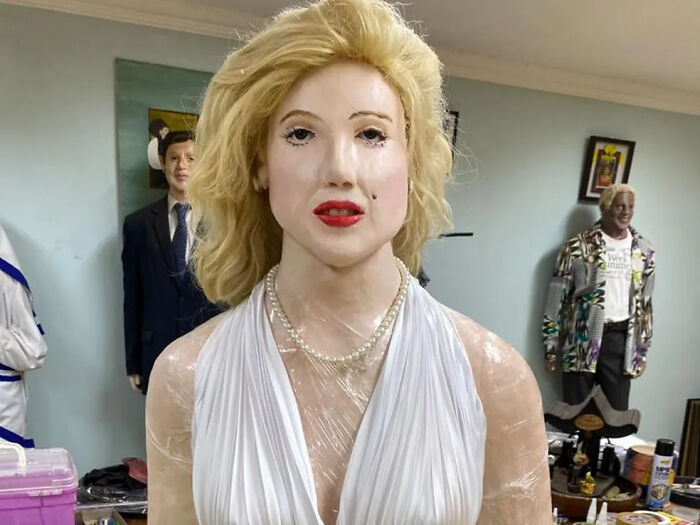 Voskovo vyzerajúca figurína s blond vlasmi v bielych šatách v miestnosti plnej dekorácií.