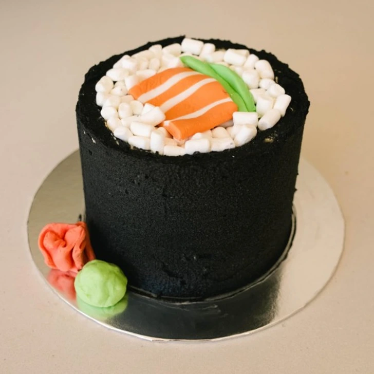 Čierno-biely tortový dezert v tvare sushi, dekorovaný lososom, zeleninou a penovými cukríkmi.
