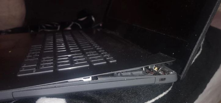 Poškodený notebook s odtrhnutým krytom. Čierna klávesnica a prasknuté pánty v detailnom zábere.