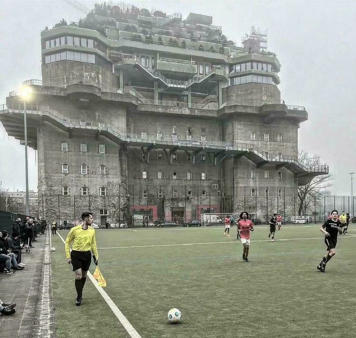 Futbalový zápas na ihrisku pri masívnej, betónovej pevnosti so zelenou strechou. Rozhodca na čiare, hráči v akcii.