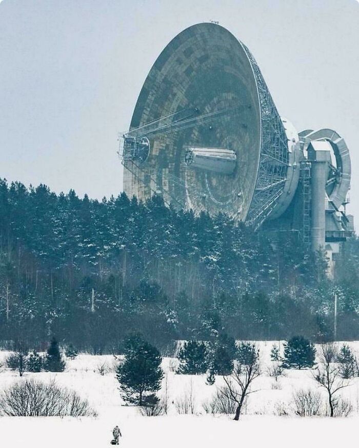 Obrovská parabolická anténa v zasneženej krajine, obklopená lesom, vyčnievajúca nad zasneženými stromami.