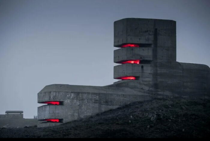 Betónová budova s červenými svetlami v tmavej krajine, vyzerá ako sci-fi pevnosť.