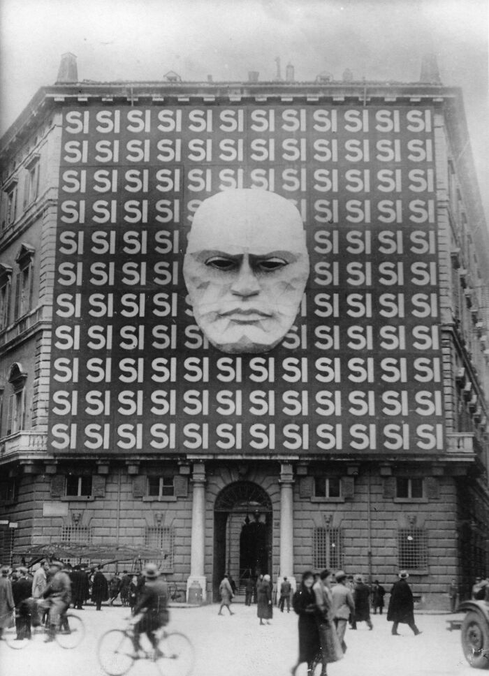Historizovaná fasáda budovy s veľkým reliéfom tváre a opakujúcimi sa nápismi SI v rušnej ulici.