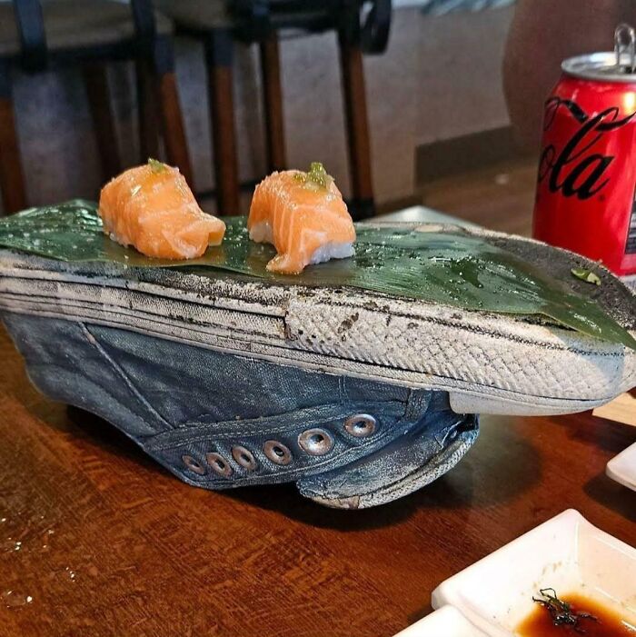 Sushi podávané netradične na starej teniske, vedľa plechovky coly na drevenom stole.