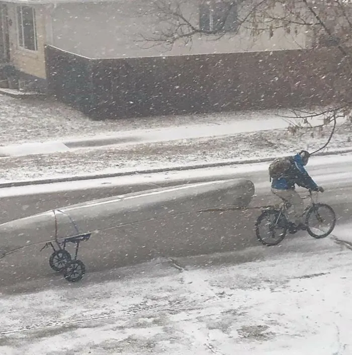 Cyklista v snežení ťahá kajak na prívesnom vozíku, prechádzajúc zimnou ulicou vedľa zasneženého domu.