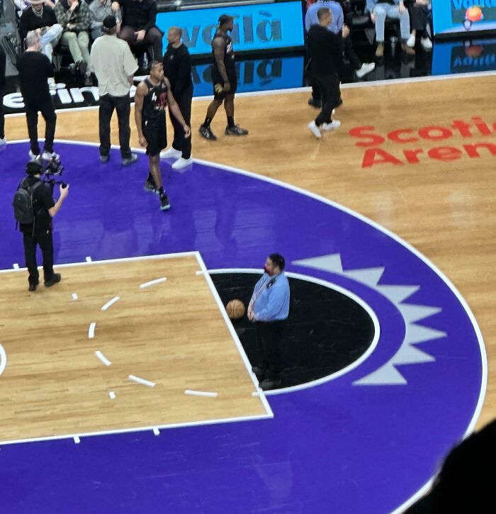 Osoba stojí na basketbalovom ihrisku s loptou, okolo prechádzajú hráči a kameraman v aréne, sledujú diváci.