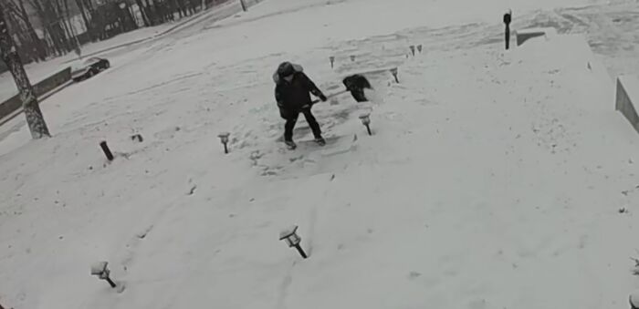Osoba odhŕňa sneh z chodníka počas silného sneženia, zasnežená scenéria s lampami a stromami v pozadí.