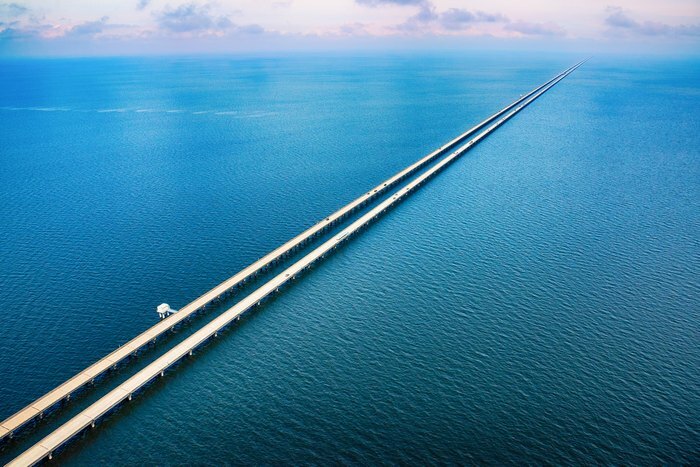 Dlhý most nad modrou vodnou hladinou za jasného dňa, tiahnuci sa až po horizont s oblohou posiatou oblakmi.