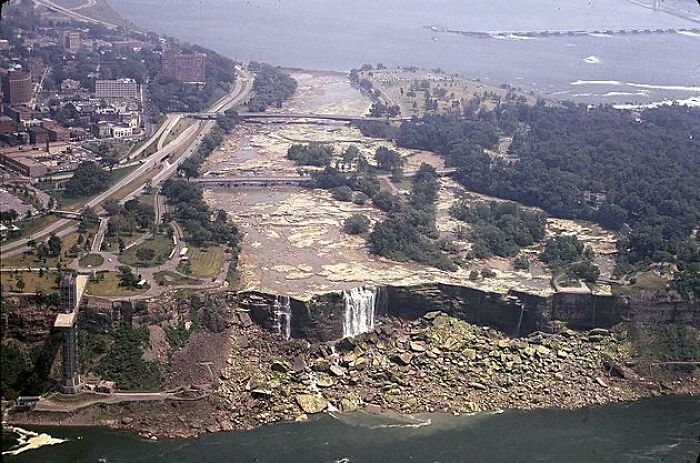 Letecký pohľad na vyschnuté Niagarské vodopády obklopené mostmi a zeleňou v pozadí veľkého mesta.