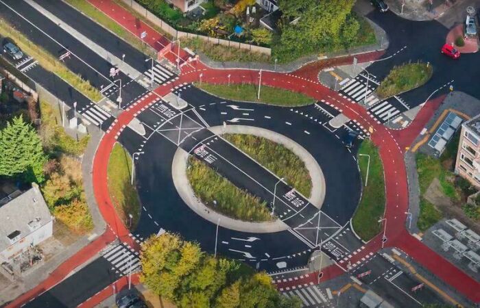 Kruhový objazd s cyklistickými pruhmi zhora, zelené oblasti, zebry pre chodcov, premávka a výhľad na okolie.