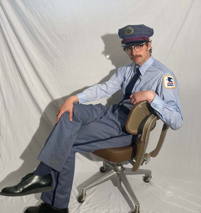 Osoba v uniforme sedí na stoličke, s klobúkom a okuliarmi, na bielom pozadí.