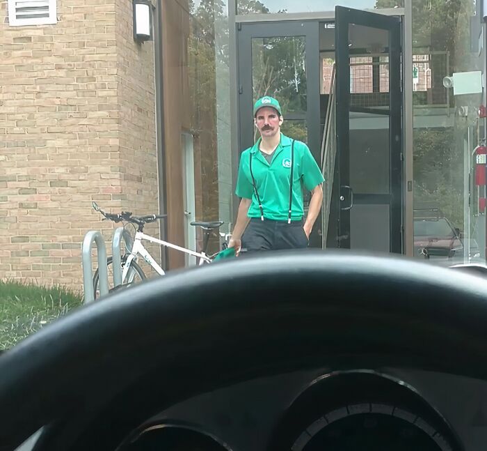 Osoba vo vychádzajúcich dverách, oblečená v zelenom oblečení, stojí vedľa bicykla pri budove.