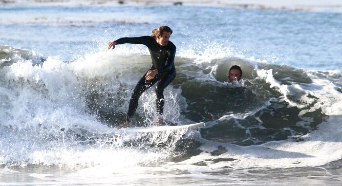 Surfer jazdí na vlne, zatiaľ čo iný človek vykúka z vody za ním na pláži.