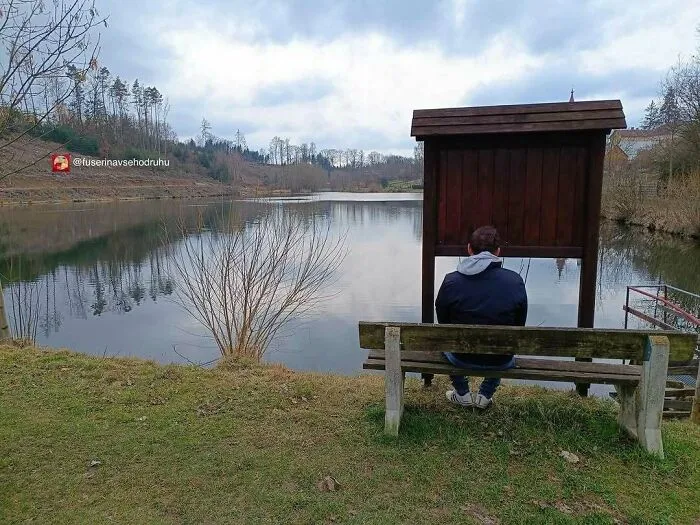 Muž sedí na lavičke pri jazere, obklopený prírodou, pod zamračenou oblohou.