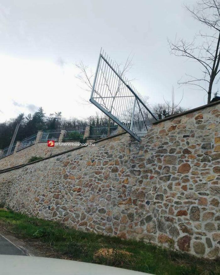 Plotová brána neštandardne postavená hore nohami na kamennom múre s výhľadom na les.