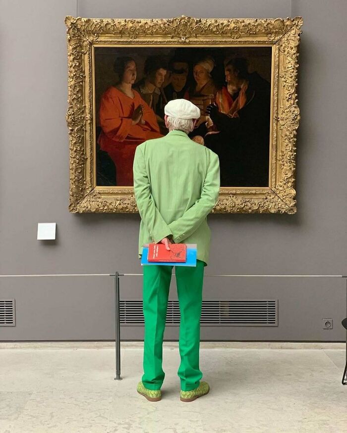 Návštevník múzea v zelenom oblečení obdivuje klasické umelecké dielo na stene galerie.