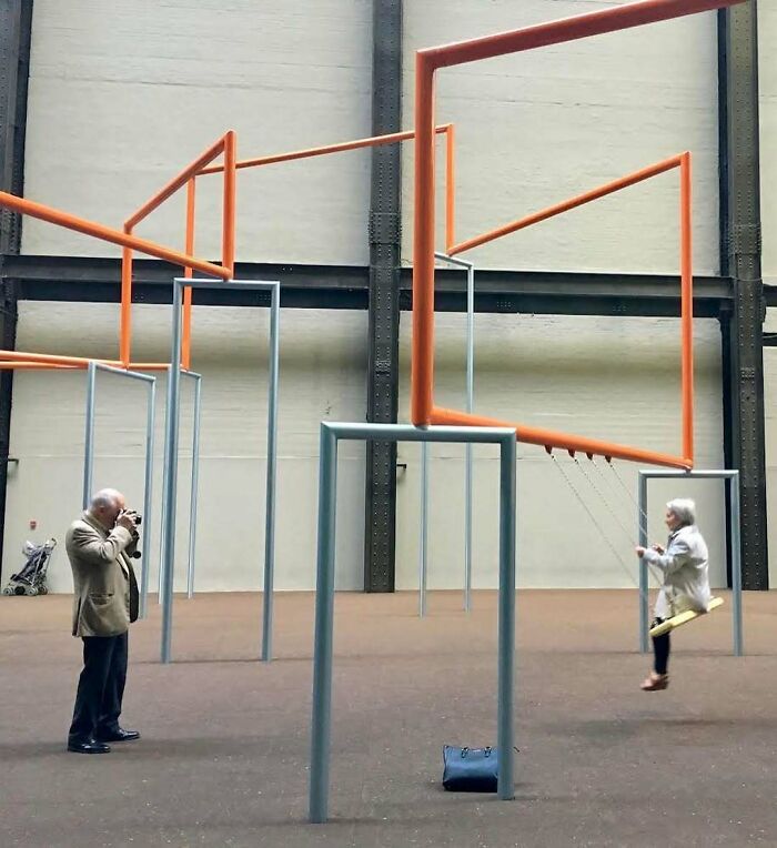 Starší muž fotografuje ženu na hojdačke v modernom interiérovom umeleckom diele s geometrickými tvarmi.