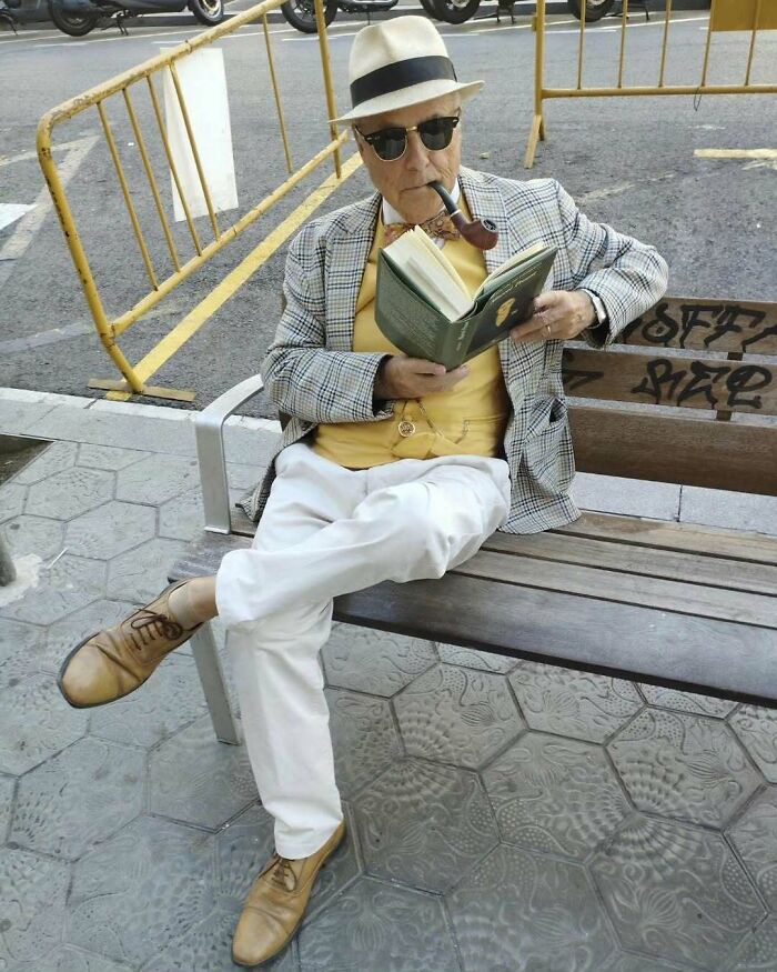 Starší pán v klobúku a fajkou číta knihu na lavičke v meste. Elegantne oblečený, uvoľnený v žltom tričku.