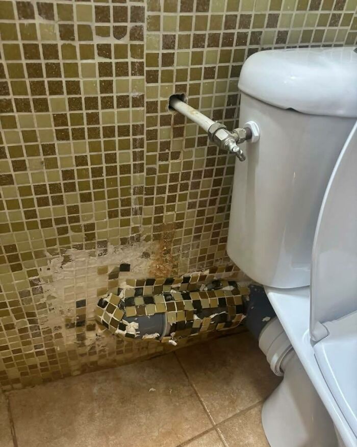 Netesniaca toaleta s poškodenou mozaikou na stene, viditeľné potrubie, potrebná oprava.