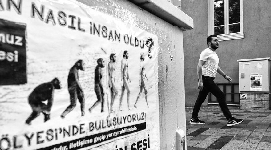 Muž kráča vedľa plagátu znázorňujúceho evolúciu človeka, čiernobiela fotografia.