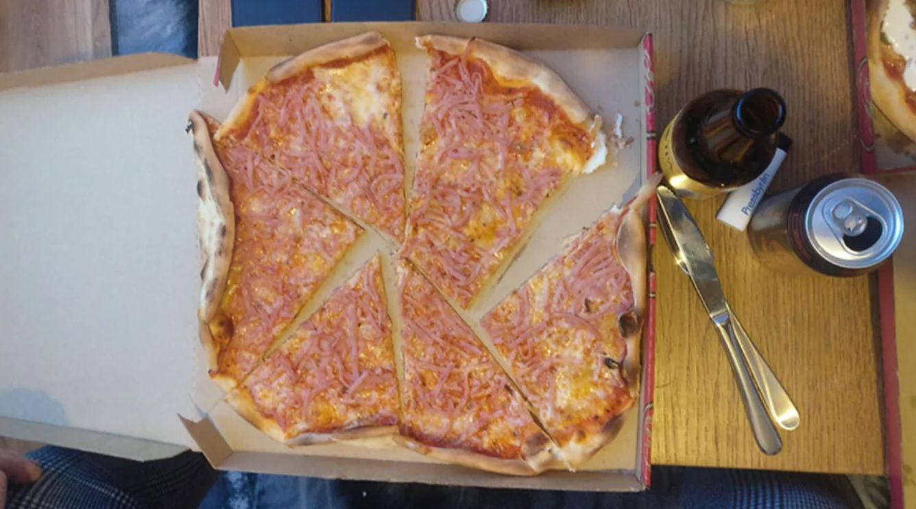 Pizza s tenkou kôrkou a šunkou na stole, obklopená nápojmi a príbormi.