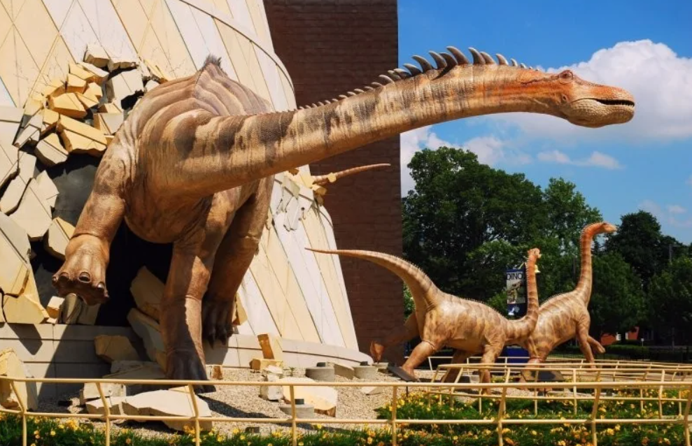 Dinosauri prerážajú múr budovy na výstave pod holým nebom, v pozadí stromy a modrá obloha.