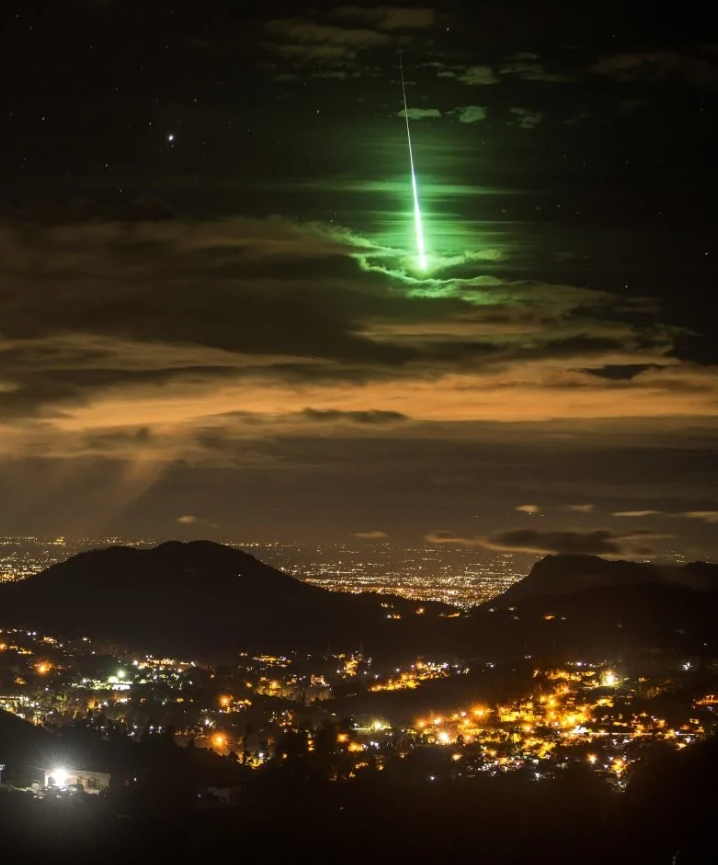 Žiarivý zelený meteor prelieta cez oblohu nad nočným mestom, osvetľujúc krajinu pod ním.
