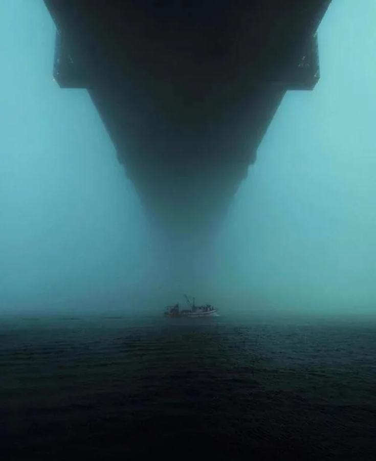 Loď pláva pod mostom v hustej hmle, ktorá vytvára tajomnú atmosféru.