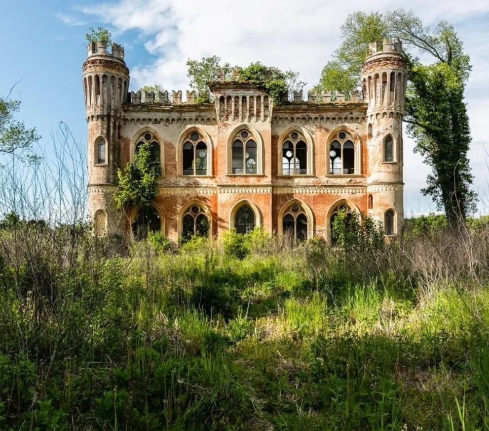 Opustený historický zámok obklopený divokou vegetáciou pod modrou oblohou.