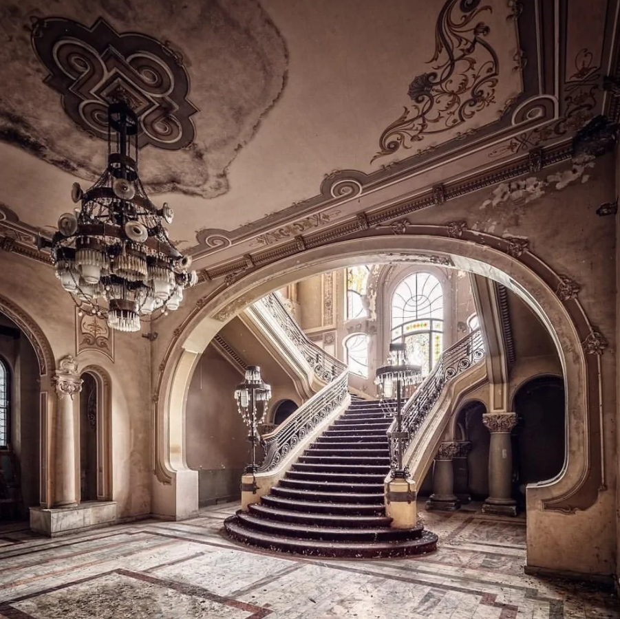 Historické schodisko v opustenom paláci s klenutým stropom a veľkým lustrom, zdôrazňujúce jeho zašlú eleganciu.
