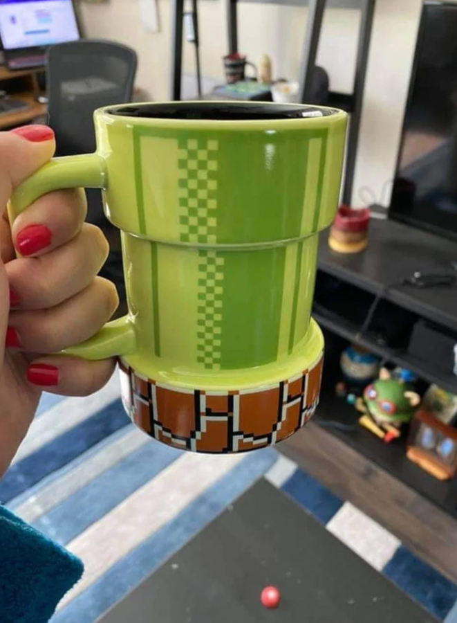 Zelený hrnček v tvare retro hernej potrubnej dekorácie držený rukou s červenými nechtami.
