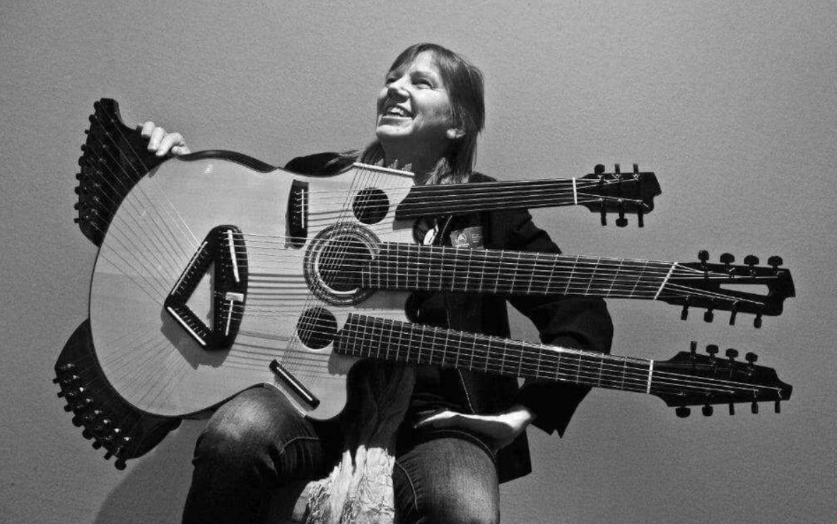 Osoba s jedinečnou gitarou s viacerými krkmi, usmievajúca sa na strohóm pozadí.