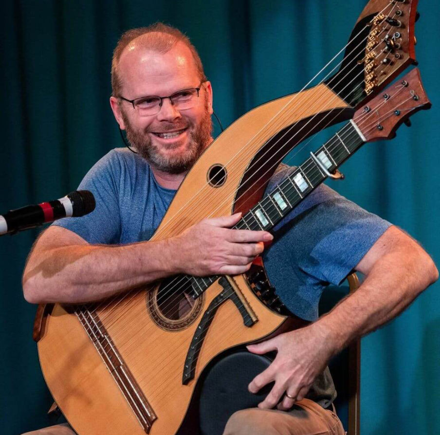 Hudobník hrá na harfovej gitare pri mikrofóne na pódiu.