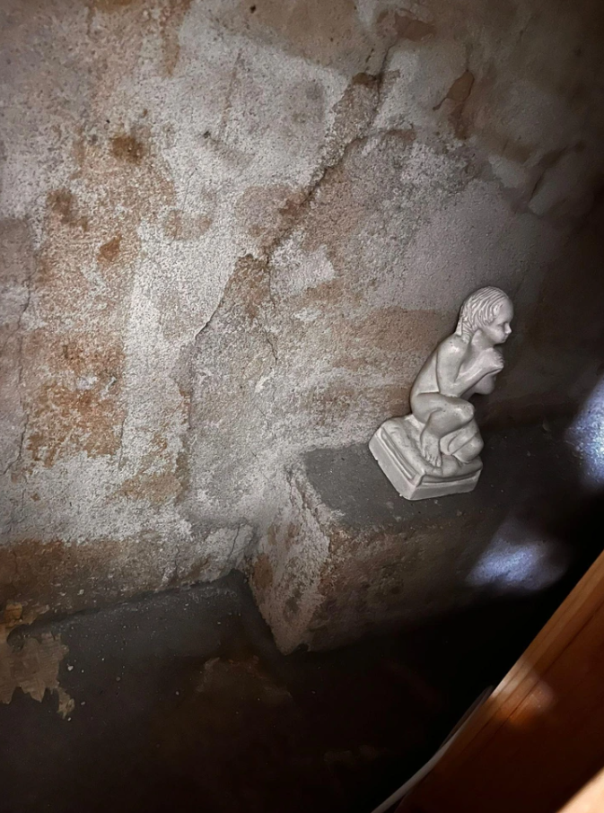 Kamenná soška sediaceho dieťaťa v tmavej miestnosti s popraskanými stenami.